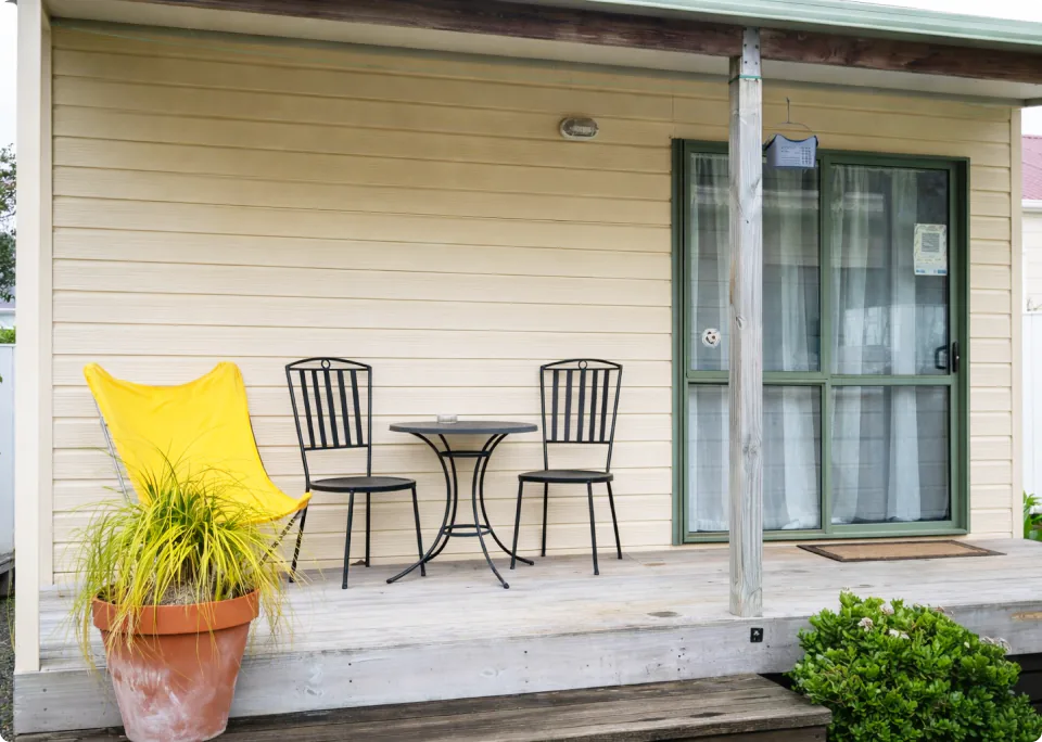 A wooden deck with black iron chairs and tables, yellow-colored chairs, and large potted plants, with a glass door allowing a view of the outdoor space inside.