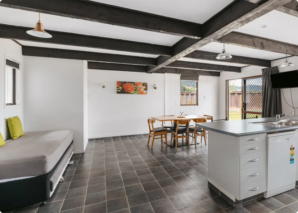 A spacious cottage interior with black beams as the distinctive ceiling, an open space with tile flooring, a bed, dining table, and kitchen counter. Large windows and glass doors allow natural light to enter, creating a cozy, comfortable room.