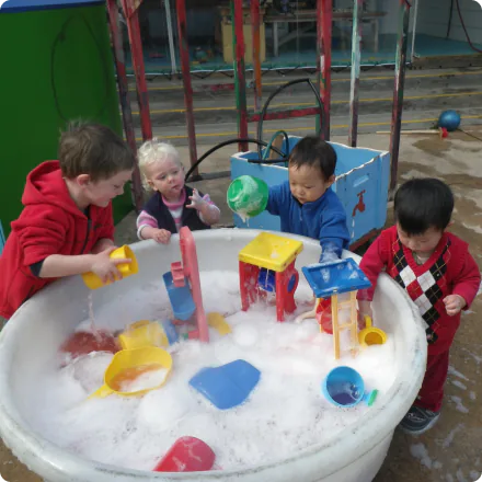 A large, bubbling aquarium filled with three toddlers and a baby child, playing with colorful scoops, toys, and water. The children are dressed in colorful clothes and enjoying the free-form water play experience typical of a play center outdoors.