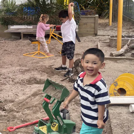A small boy in a border-pattern shirt is standing next to a green toy dump truck, smiling. A pink-dressed girl and a white-shirted boy are playing with yellow toys and shovels in a sandbox, enjoying the sunny outdoor childcare environment.