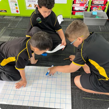 Three primary school students in black and yellow uniforms work together on a large grid paper activity in a classroom.