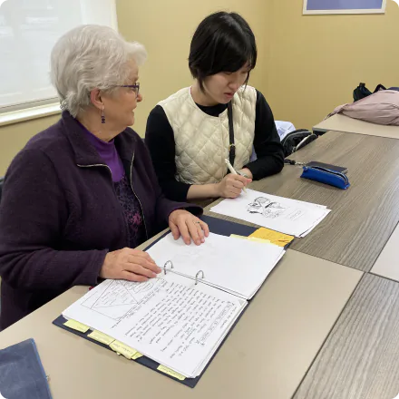 Two women, one with grey hair and a purple jacket, and the other in a best, are sitting side by side on a desk, opening English textbooks and notebooks to study together. The older woman is helping the younger woman, who is wearing a best, and they are having a conversation while looking at the notebook.