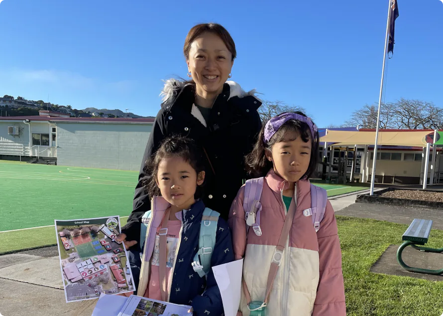 A mother and two young schoolgirls in New Zealand's schoolyard, carrying backpacks and holding school information materials. Sunny outdoor setting with school buildings and grass visible in the background.