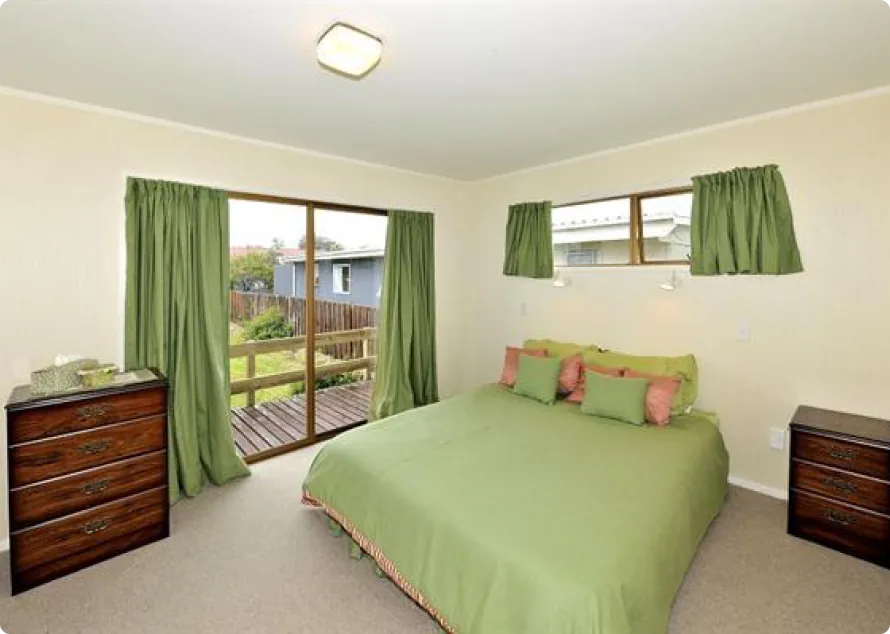 A bright bedroom in New Zealand with large windows and small windows, green curtains and bed covers, pink accent colors, wooden side tables, and a view of a wooden deck and green grass outside.