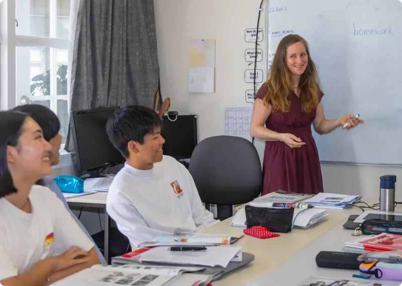 A foreign female teacher is cheerfully conducting a class using the whiteboard at the front, together with several students. The students are listening with smiles, with textbooks and notebooks spread out on the desks. The photo captures a bright, harmonious learning scene filled with natural sunlight.
