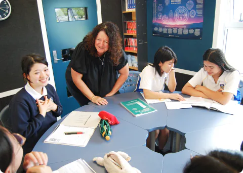 A foreign female teacher in a classroom is smiling and talking with several female students while looking at teaching materials together. The students, in uniforms, are gathered around a desk, creating a bright and lively learning environment.