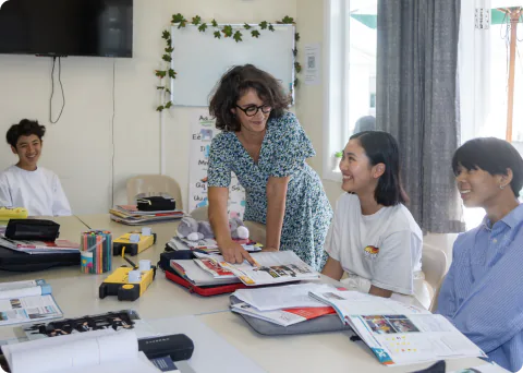 A foreign female teacher in a classroom is smiling and talking with three female students and one other student while looking at teaching materials together. The students, in uniforms, are gathered around a desk, creating a bright and lively learning environment.