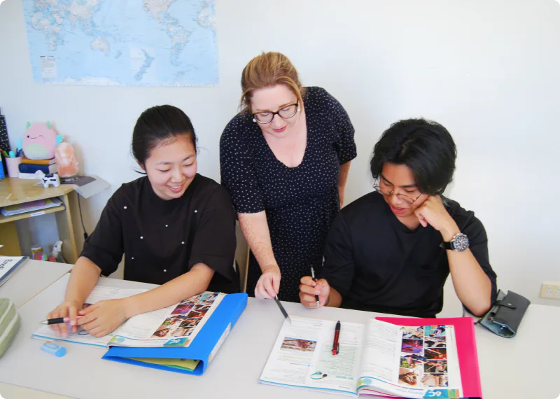 In a bright classroom, two Asian high school students are sitting at a desk with study materials spread out, while a blonde female teacher stands between them, offering attentive guidance. All three are smiling and studying in a friendly, relaxed atmosphere. A world map is displayed in the background.