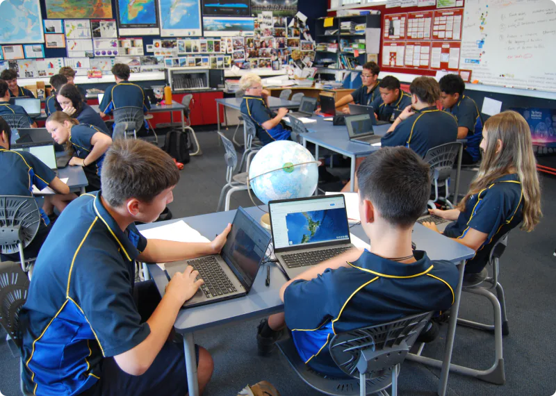 A group of high school students, wearing uniforms, are using laptops and working together on a globe and classroom displays. The students are studying in a bright, modern learning environment reminiscent of a geography class.