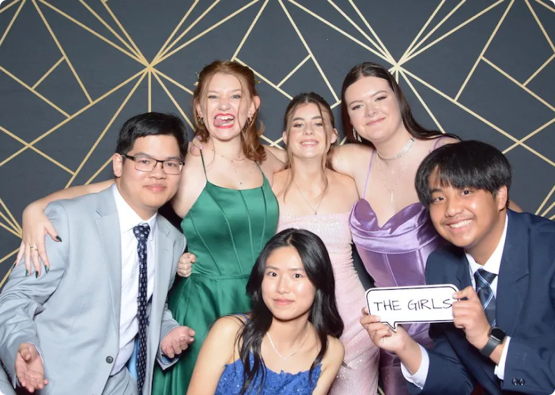A group of high school students, dressed in formal attire, gather at a party venue, smiling and holding hands while posing for the camera. The background features a geometric pattern wall. The student in the center is holding a board with the words 'THE GIRLS' written on it. A diverse group of students enjoying a friendly atmosphere and building friendships.