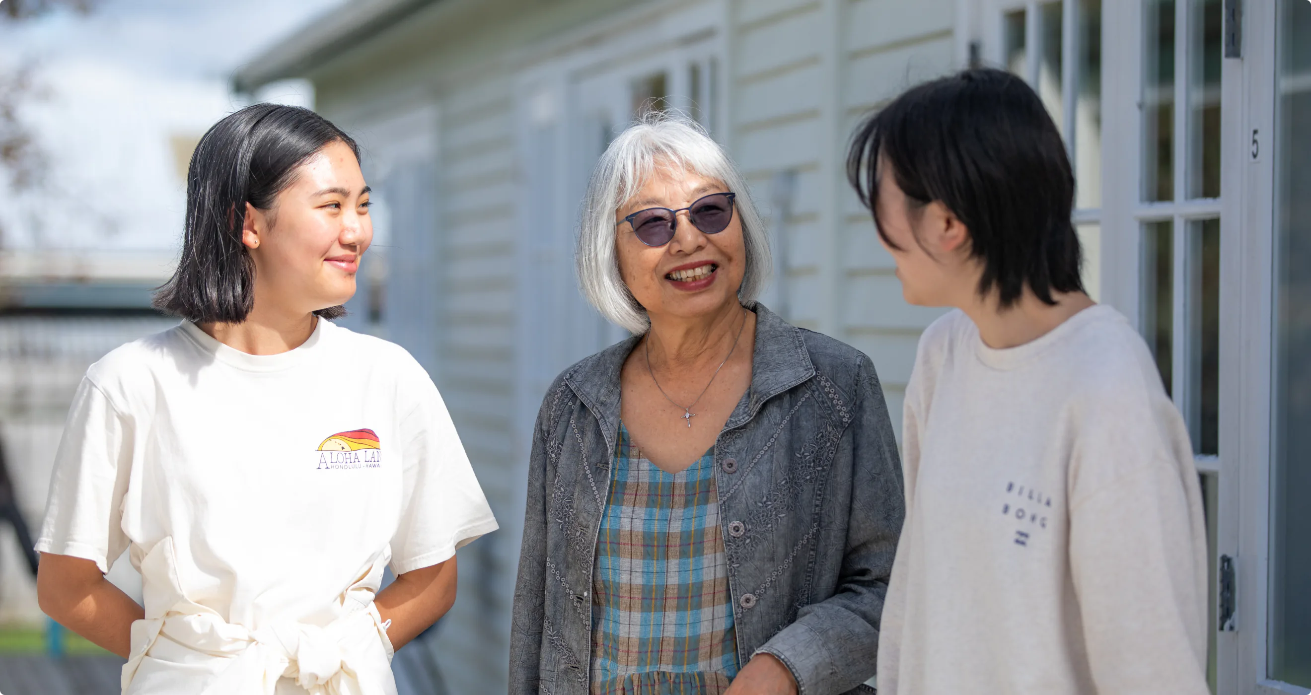 A group of young women, all wearing casual clothing, sit on a bench in an outdoor setting. A woman with gray hair is in the center, smiling and wearing sunglasses. The women are surrounded by greenery and a white wooden building in the background.